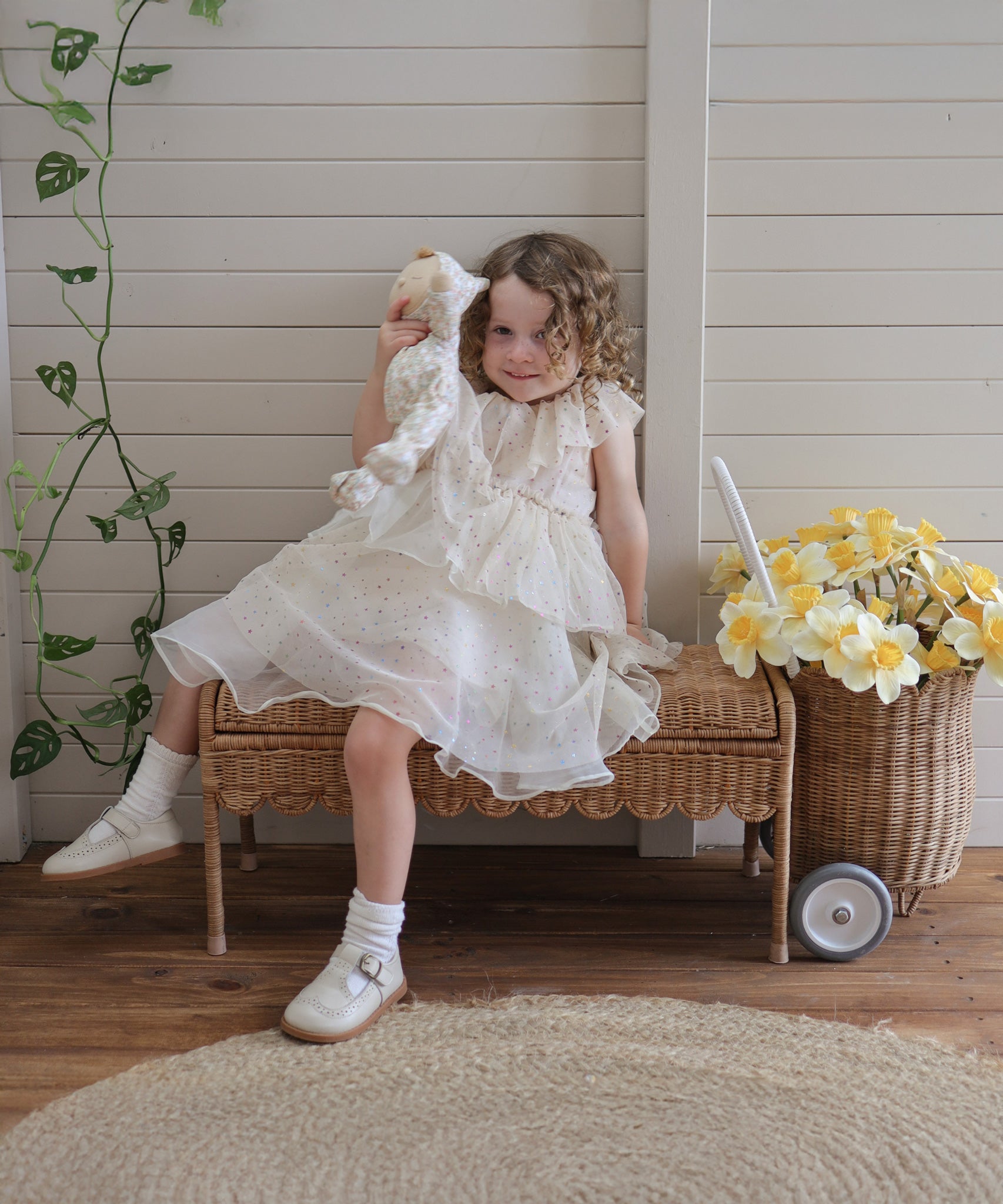 A child sitting on a Natural Rattan Olli Ella Storie Bench. The child is holding a Dozy Dinkum Doll and there is a daffodil filled petal luggy besides the bench.