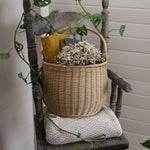 A Olli Ella Large Natural Rattan Gathering basket willed with white flowers and books placed on a wooden chair. 