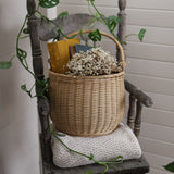 A Olli Ella Large Natural Rattan Gathering basket willed with white flowers and books placed on a wooden chair. 
