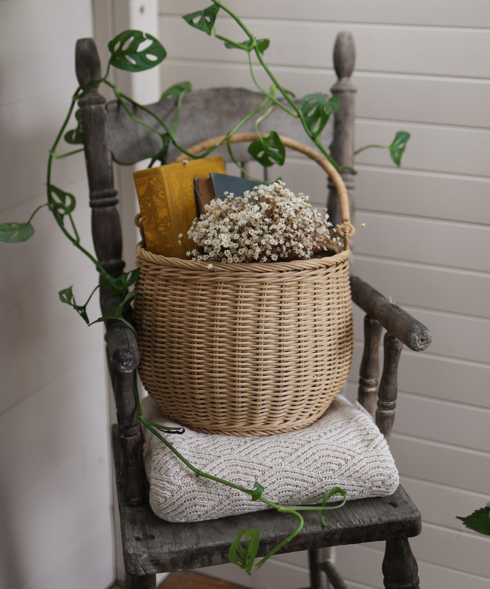 A Olli Ella Large Natural Rattan Gathering basket willed with white flowers and books placed on a wooden chair. 