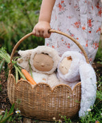 A close up of a child carrying a Natural Rattan Olli Ella Petal Basket. There are two bunny dozy dinkums and carrots inside the basket. 