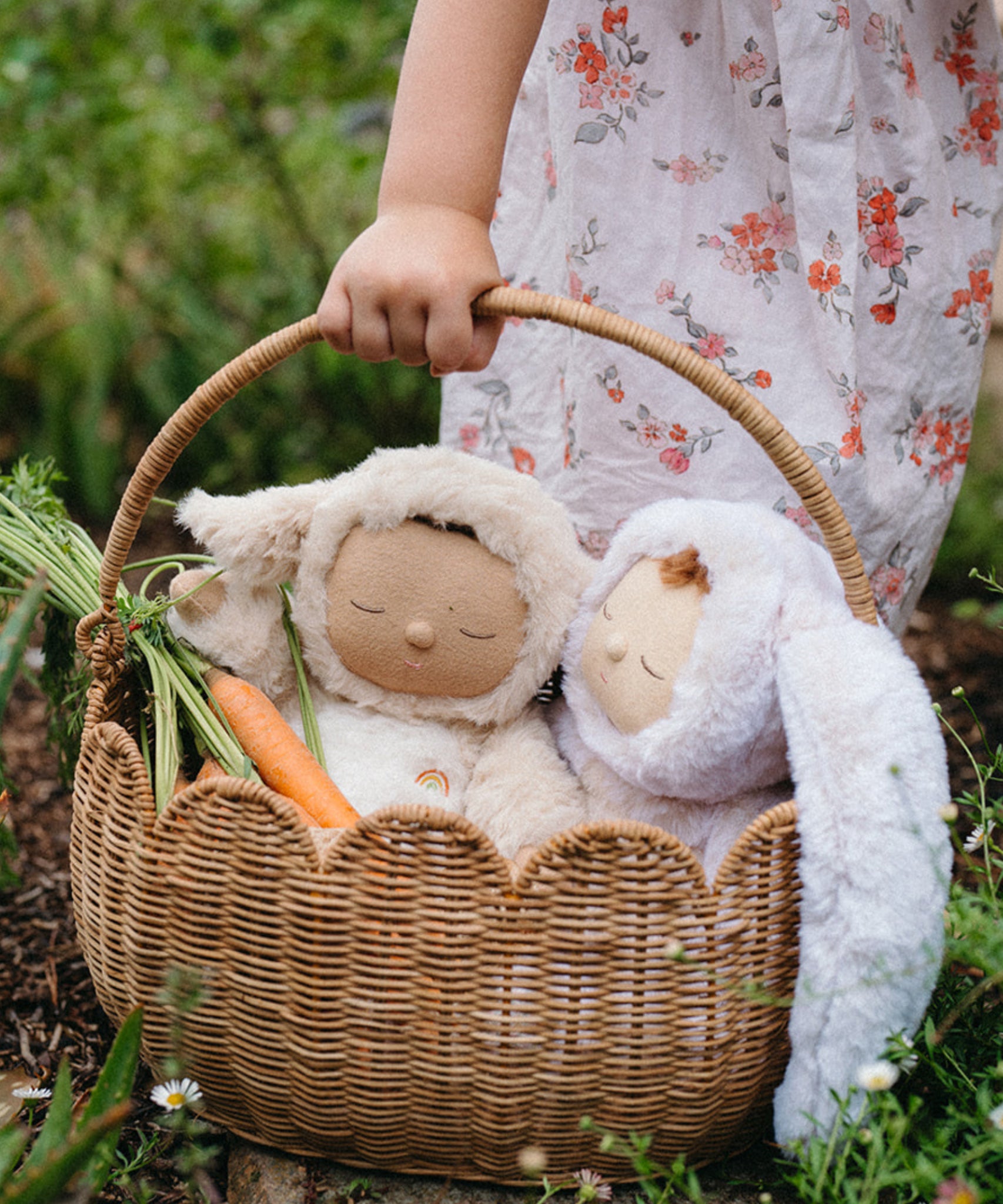 A close up of a child carrying a Natural Rattan Olli Ella Petal Basket. There are two bunny dozy dinkums and carrots inside the basket. 