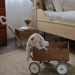 An Olli Ella Natural Rattan Petal Wonder Wagon besides a wooden bed. The Wagon has a bunny Dozy Dinkum inside. 