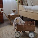 An Olli Ella Natural Rattan Petal Wonder Wagon besides a wooden bed. The Wagon has a bunny Dozy Dinkum inside. 