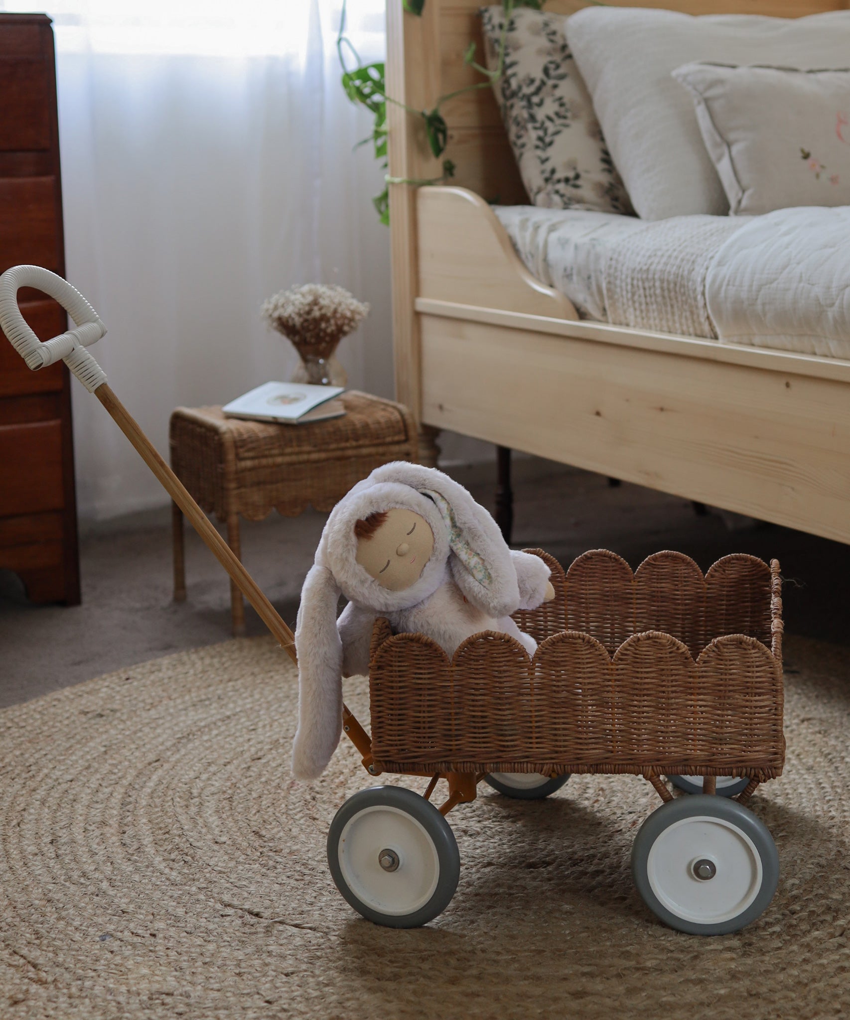 An Olli Ella Natural Rattan Petal Wonder Wagon besides a wooden bed. The Wagon has a bunny Dozy Dinkum inside. 