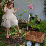 A child in a dress sitting on a swing. They have a Natural Rattan Olli Ella Petal Wonder Wagon on the ground besides them.