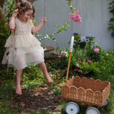 A child in a dress sitting on a swing. They have a Natural Rattan Olli Ella Petal Wonder Wagon on the ground besides them.