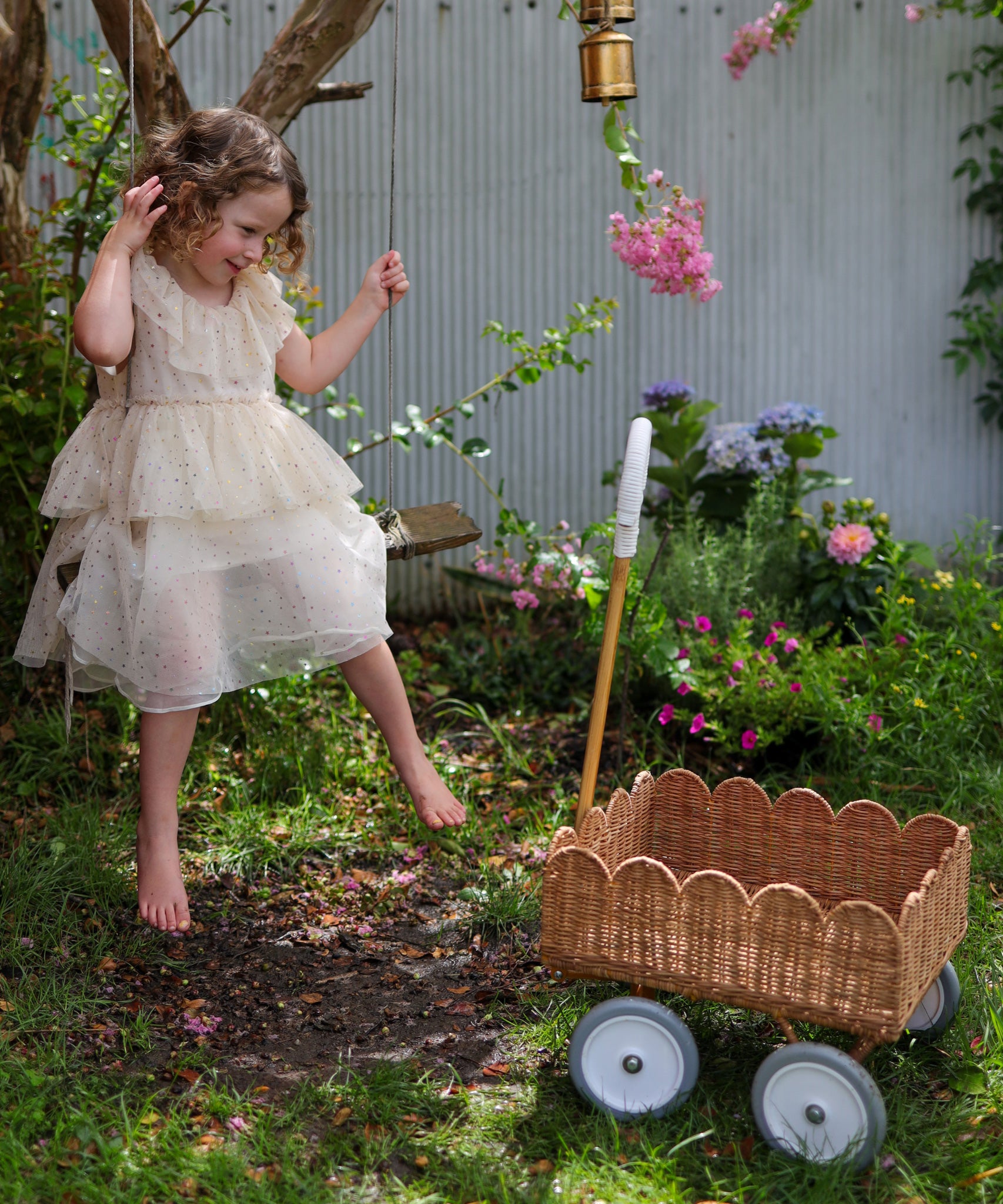 A child in a dress sitting on a swing. They have a Natural Rattan Olli Ella Petal Wonder Wagon on the ground besides them.