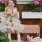 A child sitting on a Seashell Pink Olli Ella Storie Bench. The child also has a Dinkum Doll in their arms.