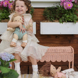 A child sitting on a Seashell Pink Olli Ella Storie Bench. The child also has a Dinkum Doll in their arms.