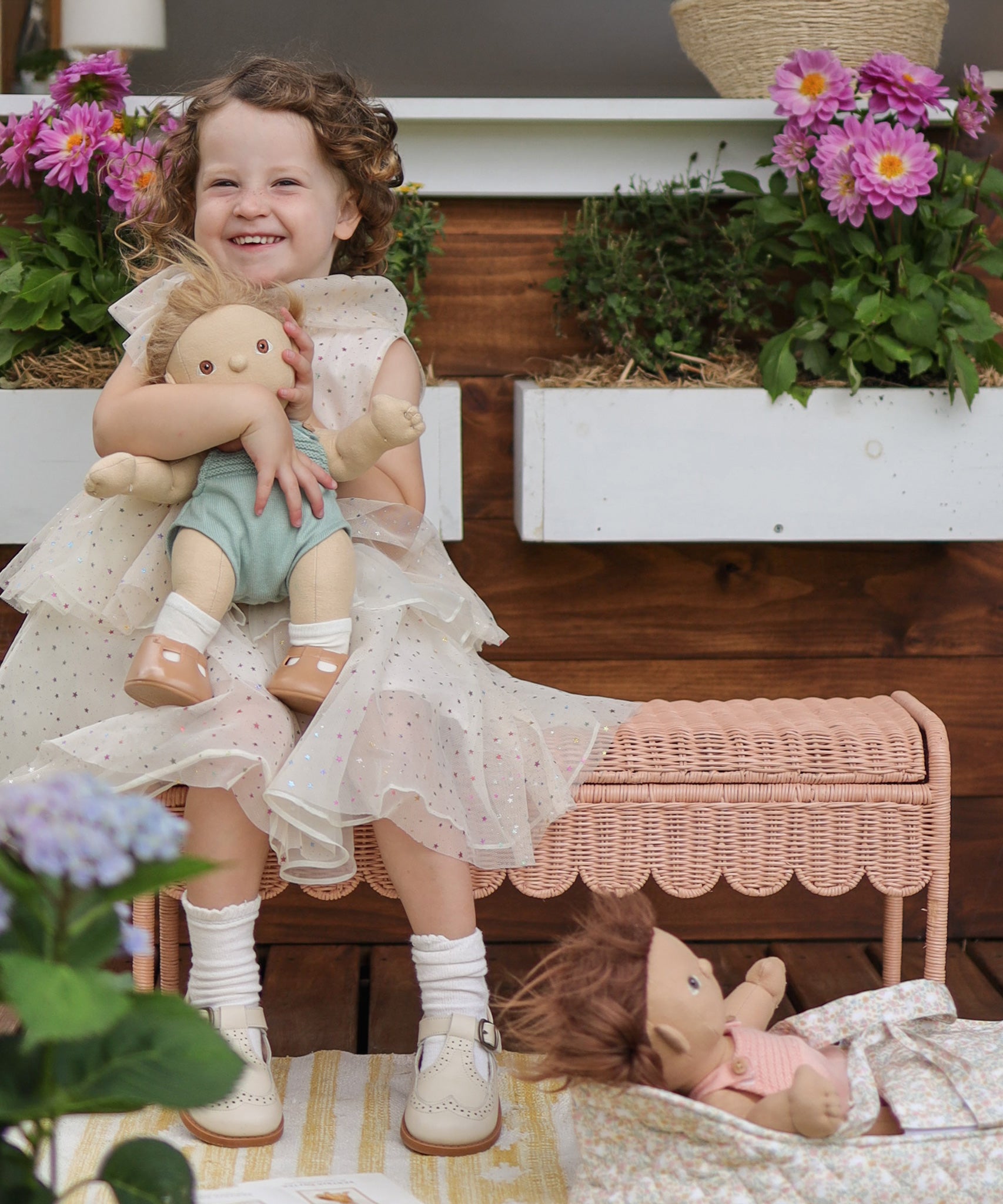 A child sitting on a Seashell Pink Olli Ella Storie Bench. The child also has a Dinkum Doll in their arms.