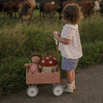A child pushing a Seashell Pink Olli Ella Petal Wonder Wagon along in front of them. The wagon has a Dinkum Doll and Ollie Ella toadstool basket inside. 