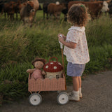 A child pushing a Seashell Pink Olli Ella Petal Wonder Wagon along in front of them. The wagon has a Dinkum Doll and Ollie Ella toadstool basket inside. 