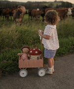 A child pushing a Seashell Pink Olli Ella Petal Wonder Wagon along in front of them. The wagon has a Dinkum Doll and Ollie Ella toadstool basket inside. 