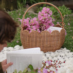 A Olli Ella Natural Rattan Petal Basket filled with pink flowers and a book. A child can be seen to the side reading a book on a blanket. 