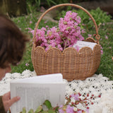 A Olli Ella Natural Rattan Petal Basket filled with pink flowers and a book. A child can be seen to the side reading a book on a blanket. 