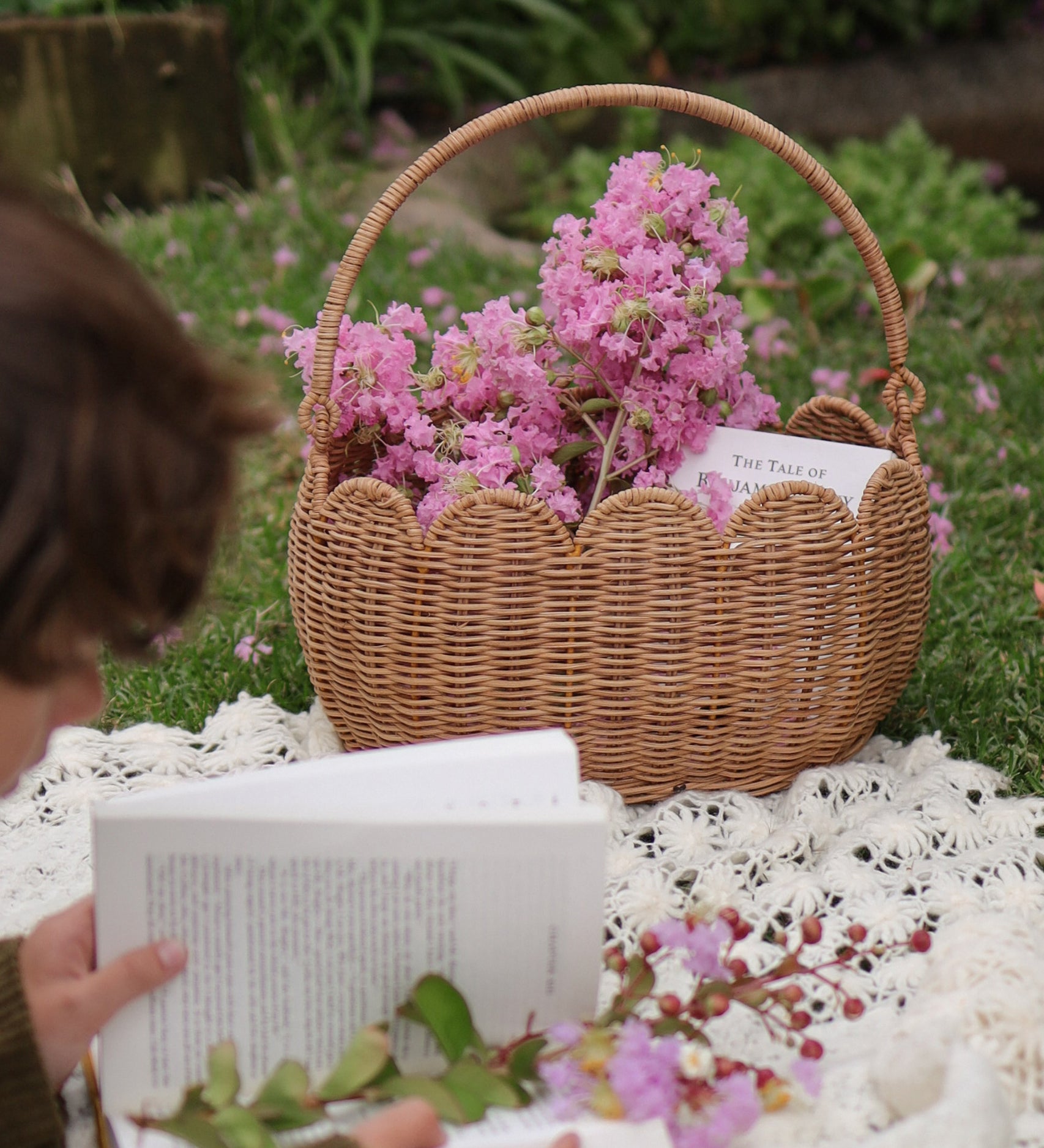 A Olli Ella Natural Rattan Petal Basket filled with pink flowers and a book. A child can be seen to the side reading a book on a blanket. 