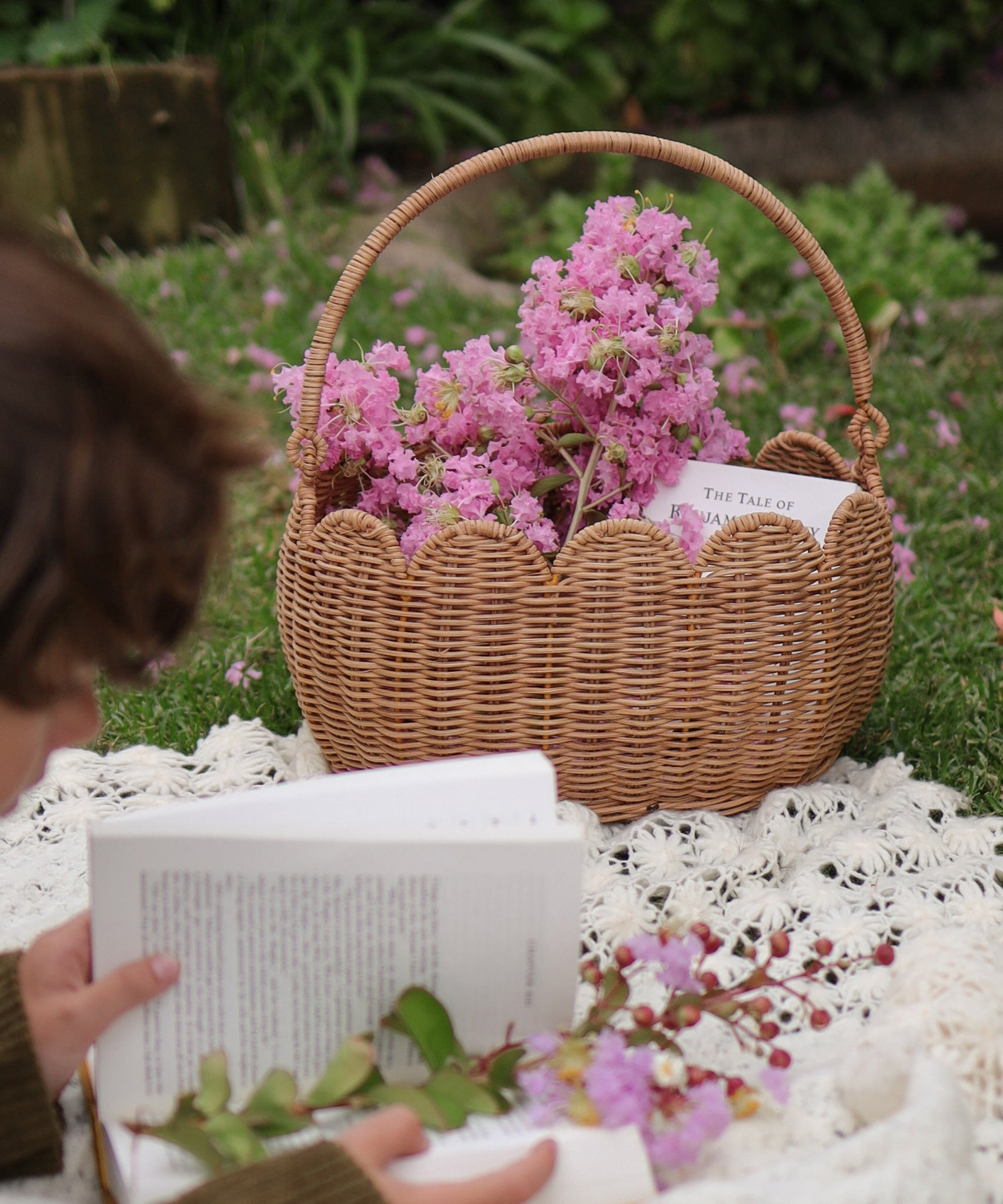 A Olli Ella Natural Rattan Petal Basket filled with pink flowers and a book. A child can be seen to the side reading a book on a blanket. 