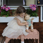 A child sitting on a Seashell Pink Olli Ella Storie Bench. The child also has a Dinkum Doll in their arms which they are holding besides them.