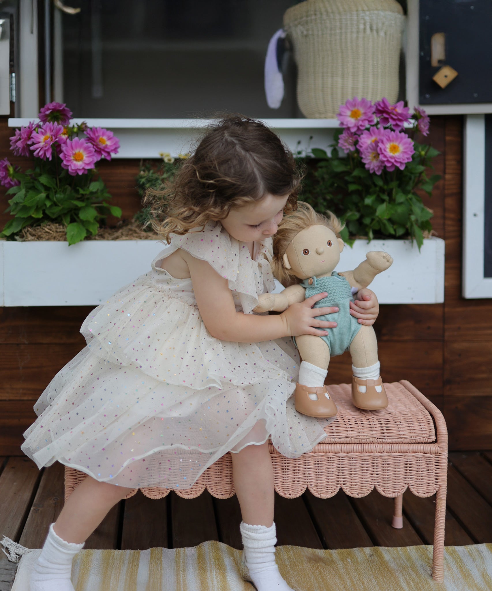 A child sitting on a Seashell Pink Olli Ella Storie Bench. The child also has a Dinkum Doll in their arms which they are holding besides them.