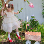 A child in a dress sitting on a swing. They have a Seashell Pink Olli Ella Petal Wonder Wagon on the ground besides them.