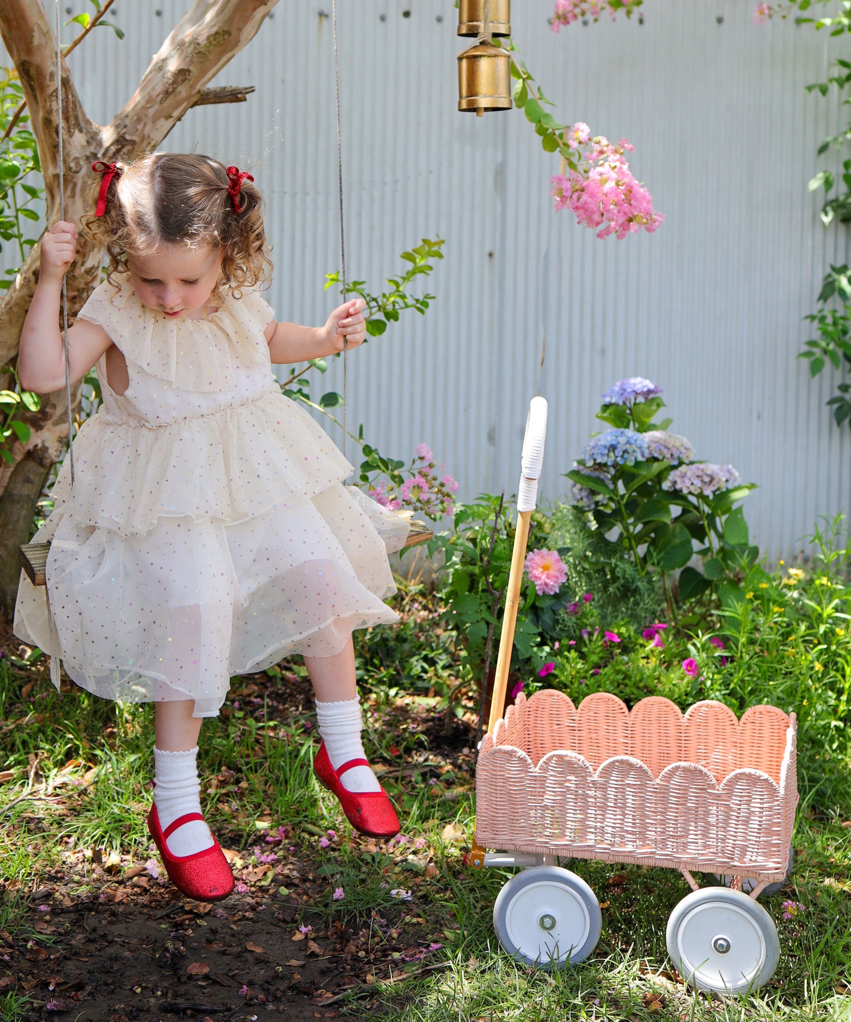 A child in a dress sitting on a swing. They have a Seashell Pink Olli Ella Petal Wonder Wagon on the ground besides them.