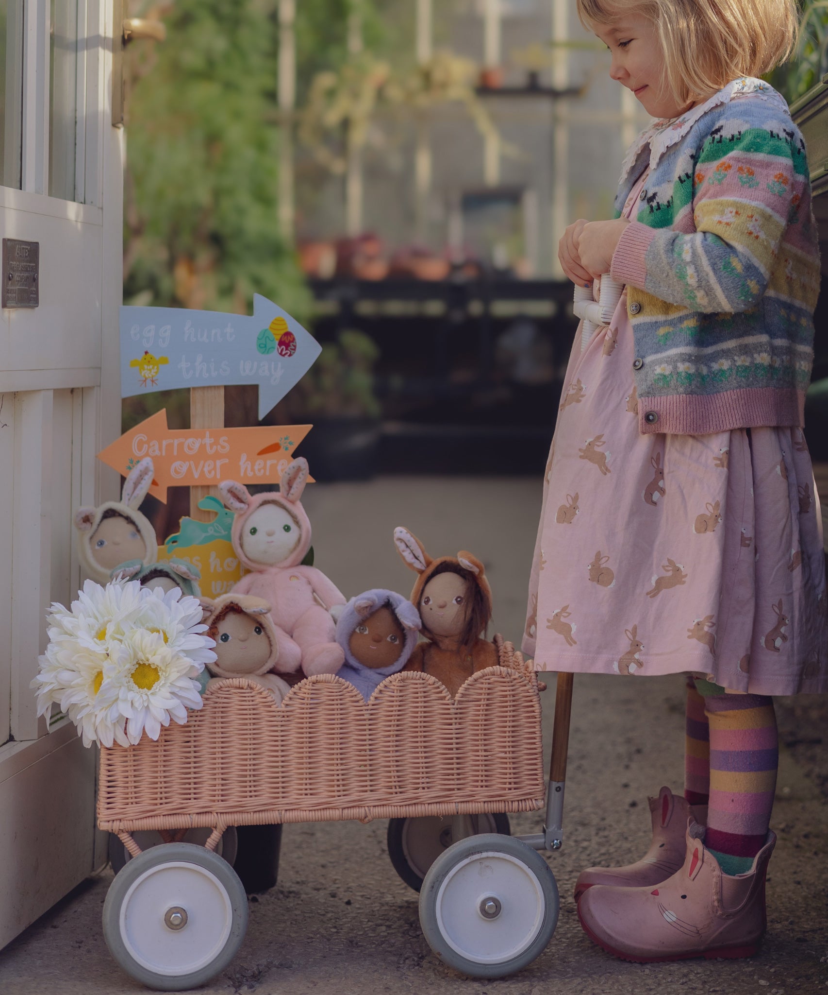A child pushing a Seashell Pink Olli Ella Petal Wonder Wagon along in front of them. The wagon is filled with Dinky Dinkums and a bunch of white flowers. 