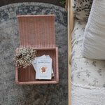 Olli Ella Petal Storie Stool in Seashell Pink Rattan. The stool has been placed by the side of a bed and has books placed inside, the lid has been left open.