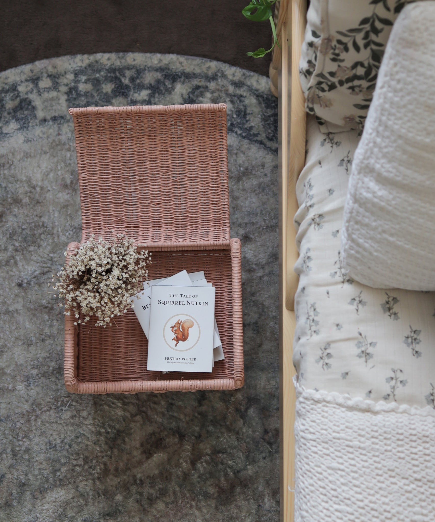 Olli Ella Petal Storie Stool in Seashell Pink Rattan. The stool has been placed by the side of a bed and has books placed inside, the lid has been left open.