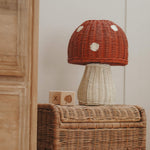 The Olli Ella rattan toadstool lamp with a white base and a red mushroom top, on a rattan table. 