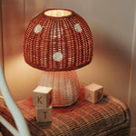 The Olli Ella rattan toadstool lamp with a white base and a red mushroom top, on a toy box and creating oft warm light.
