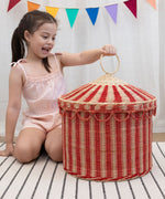 A child lifting the top off the Olli Ella Red & Straw Rattan Circus Tent Basket. This large capacity basket is perfect for use as toy storage. Olli Ella have a range of stylish storage solutions and soft toys available here at Babipur. 