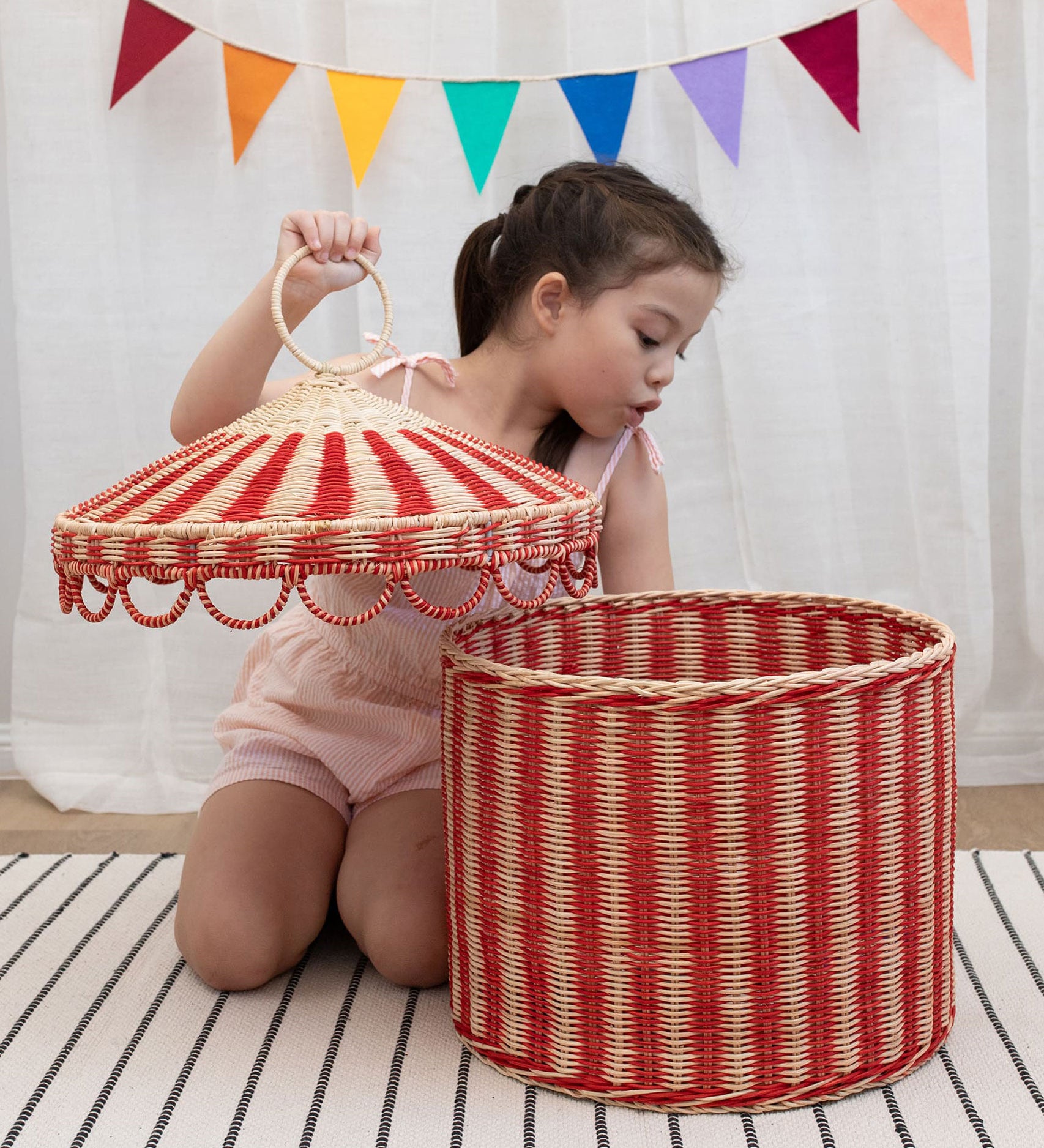 A child lifting the lid off the Olli Ella Red & Straw Rattan Circus Tent Basket. This large capacity basket is perfect for use as toy storage. Olli Ella have a range of stylish storage solutions and soft toys available here at Babipur. 