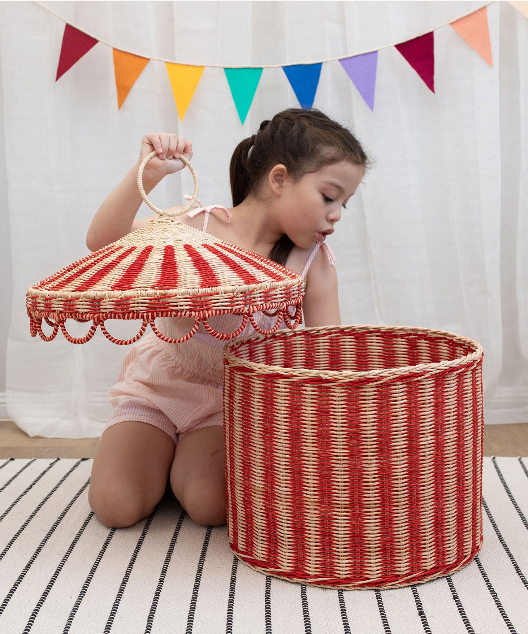 A child lifting the lid off the Olli Ella Red & Straw Rattan Circus Tent Basket. This large capacity basket is perfect for use as toy storage. Olli Ella have a range of stylish storage solutions and soft toys available here at Babipur. 