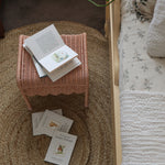Olli Ella Petal Storie Stool in Seashell Pink Rattan. The stool is shown next to a bed with books placed on top