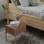 Olli Ella Petal Storie Stool in Seashell Pink Rattan. The stool has been placed by the side of a bed and has books and a small vase of white flowers placed on top.