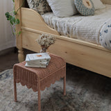 Olli Ella Petal Storie Stool in Seashell Pink Rattan. The stool has been placed by the side of a bed and has books and a small vase of white flowers placed on top.