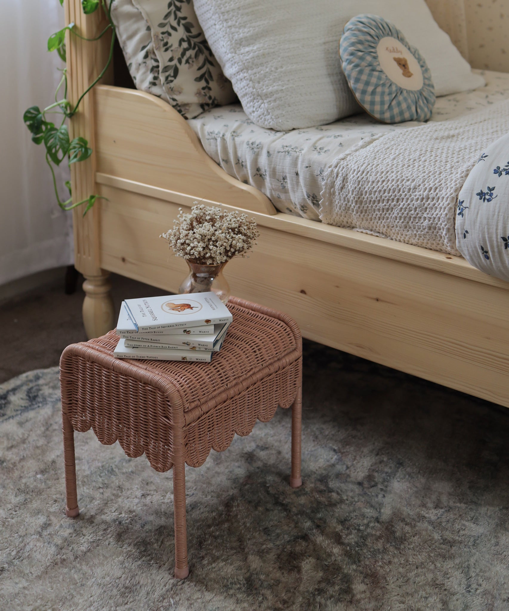 Olli Ella Petal Storie Stool in Seashell Pink Rattan. The stool has been placed by the side of a bed and has books and a small vase of white flowers placed on top.
