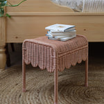 Olli Ella Petal Storie Stool in Seashell Pink Rattan. The stool has been placed by the side of a wooden bed and has books piled on the top. 