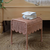 Olli Ella Petal Storie Stool in Seashell Pink Rattan. The stool has been placed by the side of a wooden bed and has books piled on the top. 