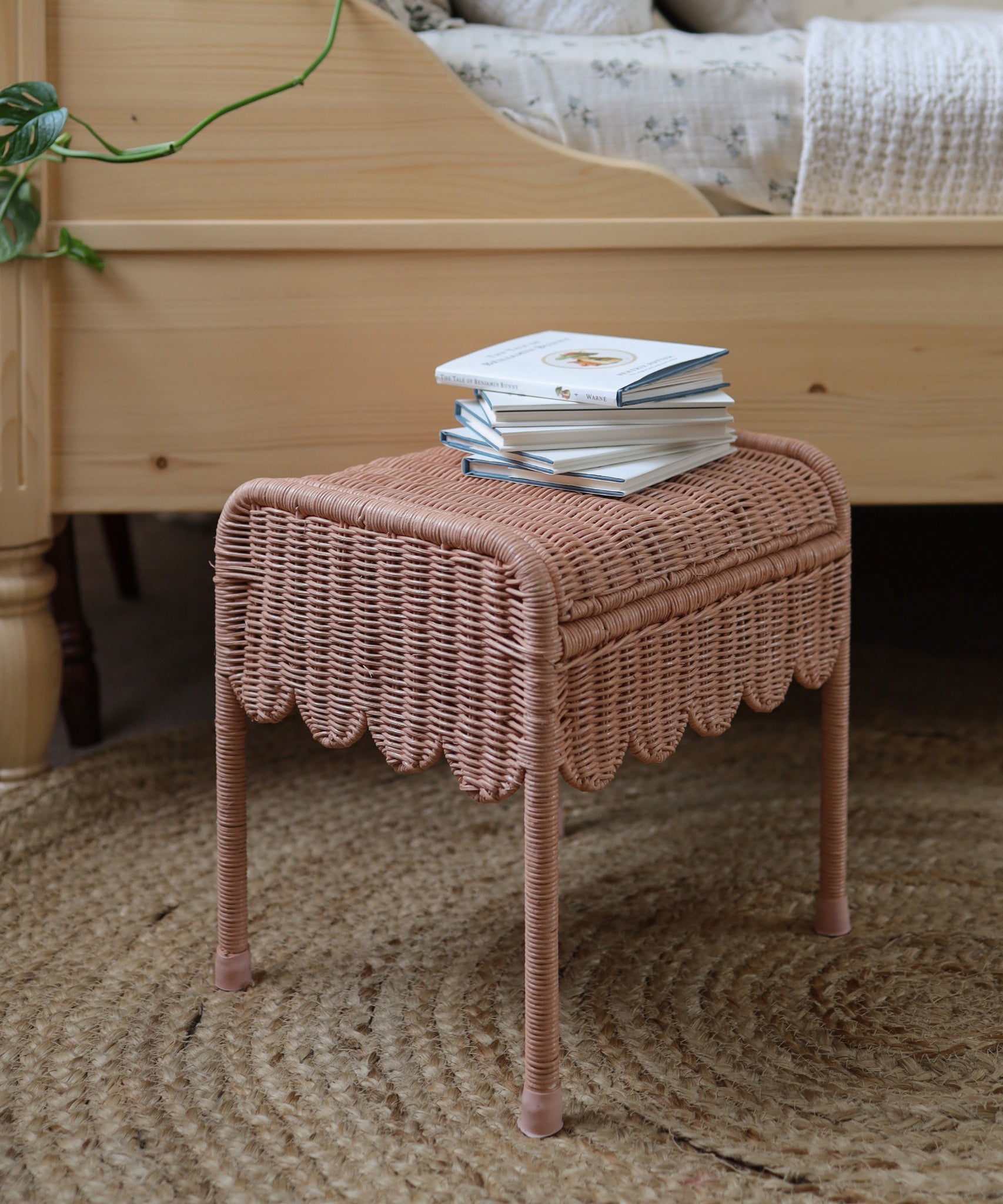 Olli Ella Petal Storie Stool in Seashell Pink Rattan. The stool has been placed by the side of a wooden bed and has books piled on the top. 