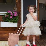 A child standing outdoors in a party dress holding a seashell pink Olli Ella Rattan Petal Luggy by their side.