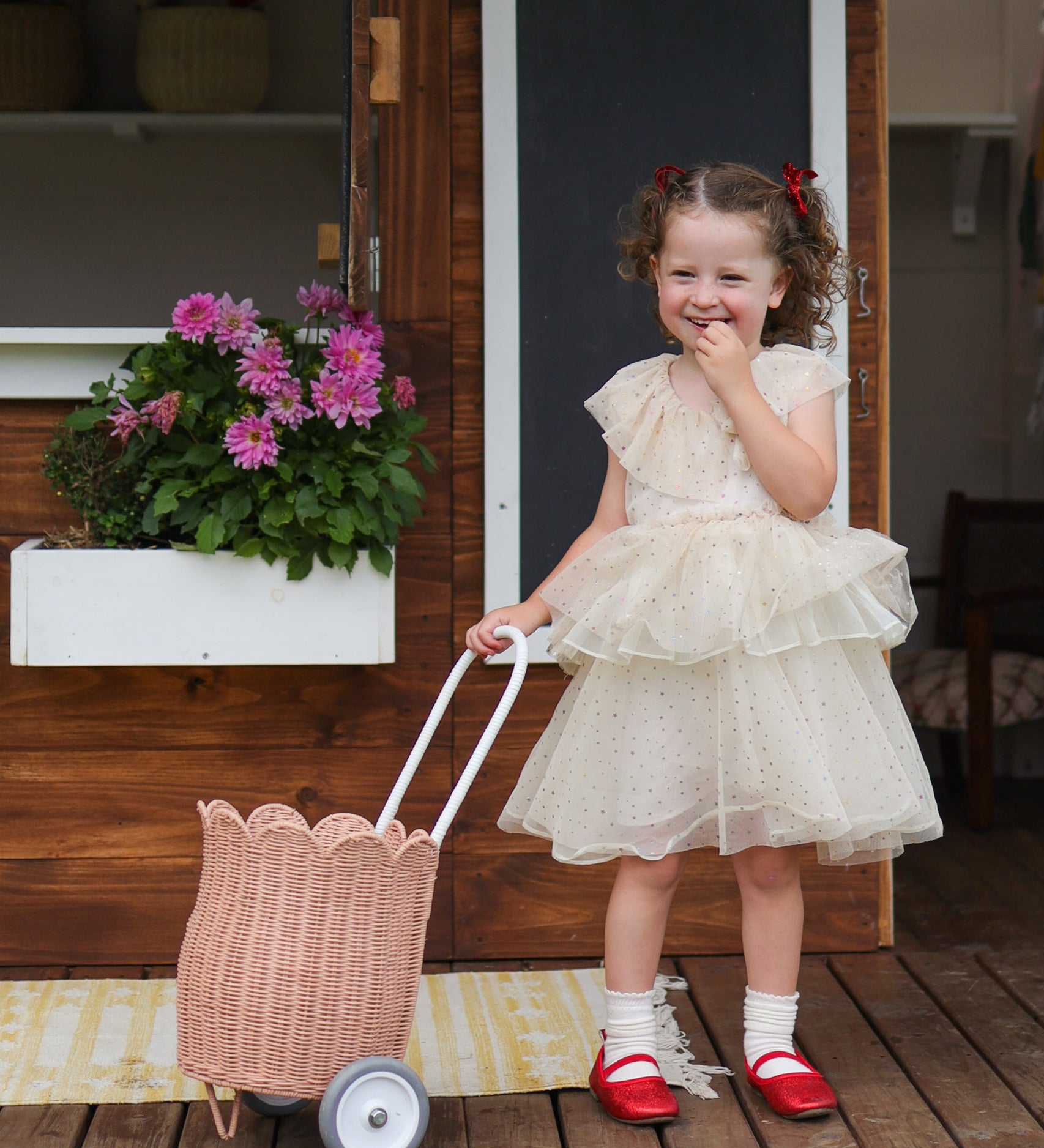 A child standing outdoors in a party dress holding a seashell pink Olli Ella Rattan Petal Luggy by their side.