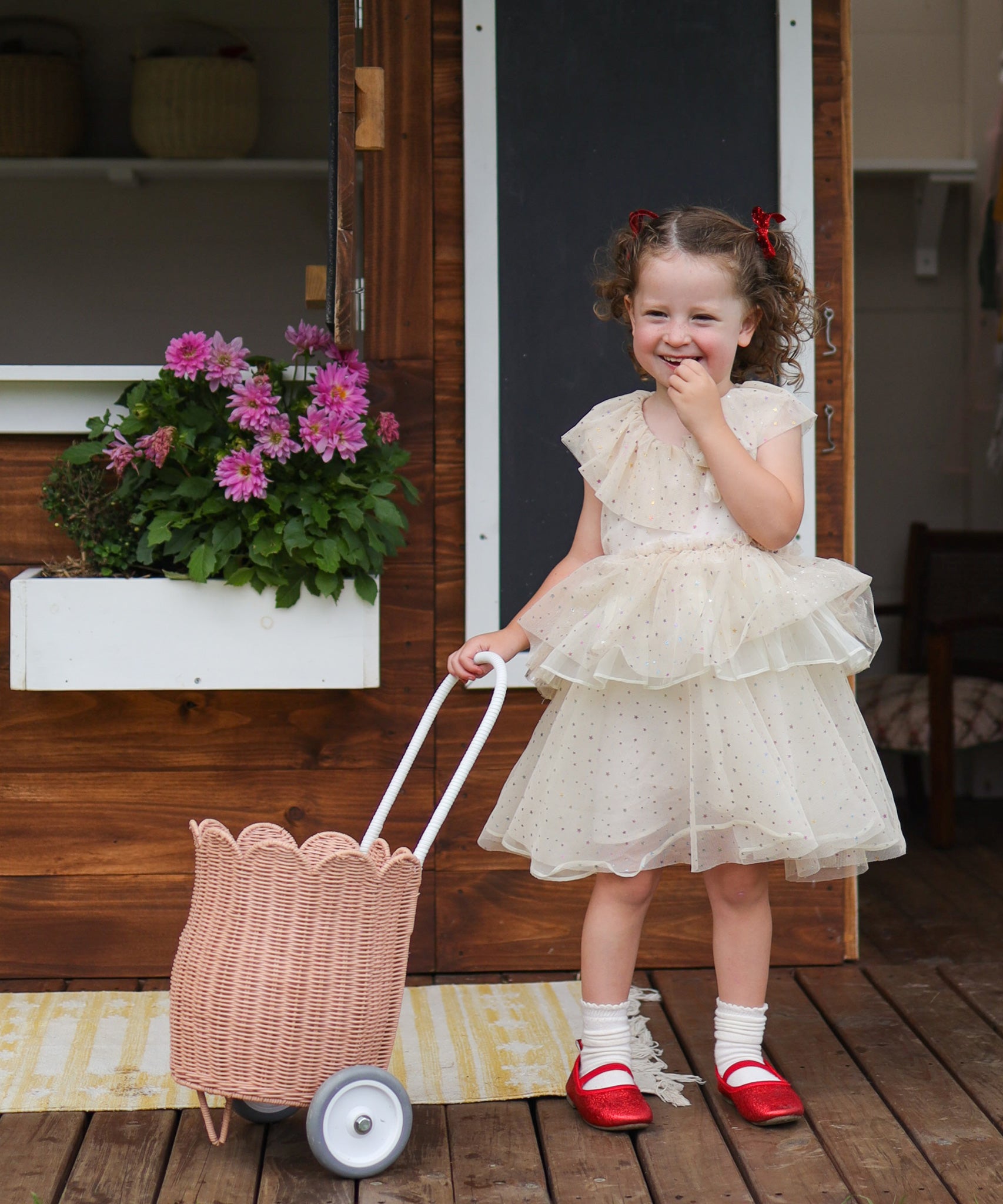 A child standing outdoors in a party dress holding a seashell pink Olli Ella Rattan Petal Luggy by their side.