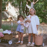 Two children standing outdoors. One is wearing the Olli Ella bunny ears headband and has a Seashell Pink rattan petal luggy by their side. Both children are holding Dozy Dinkum soft toys. There is also a rattan wagon off to the side.