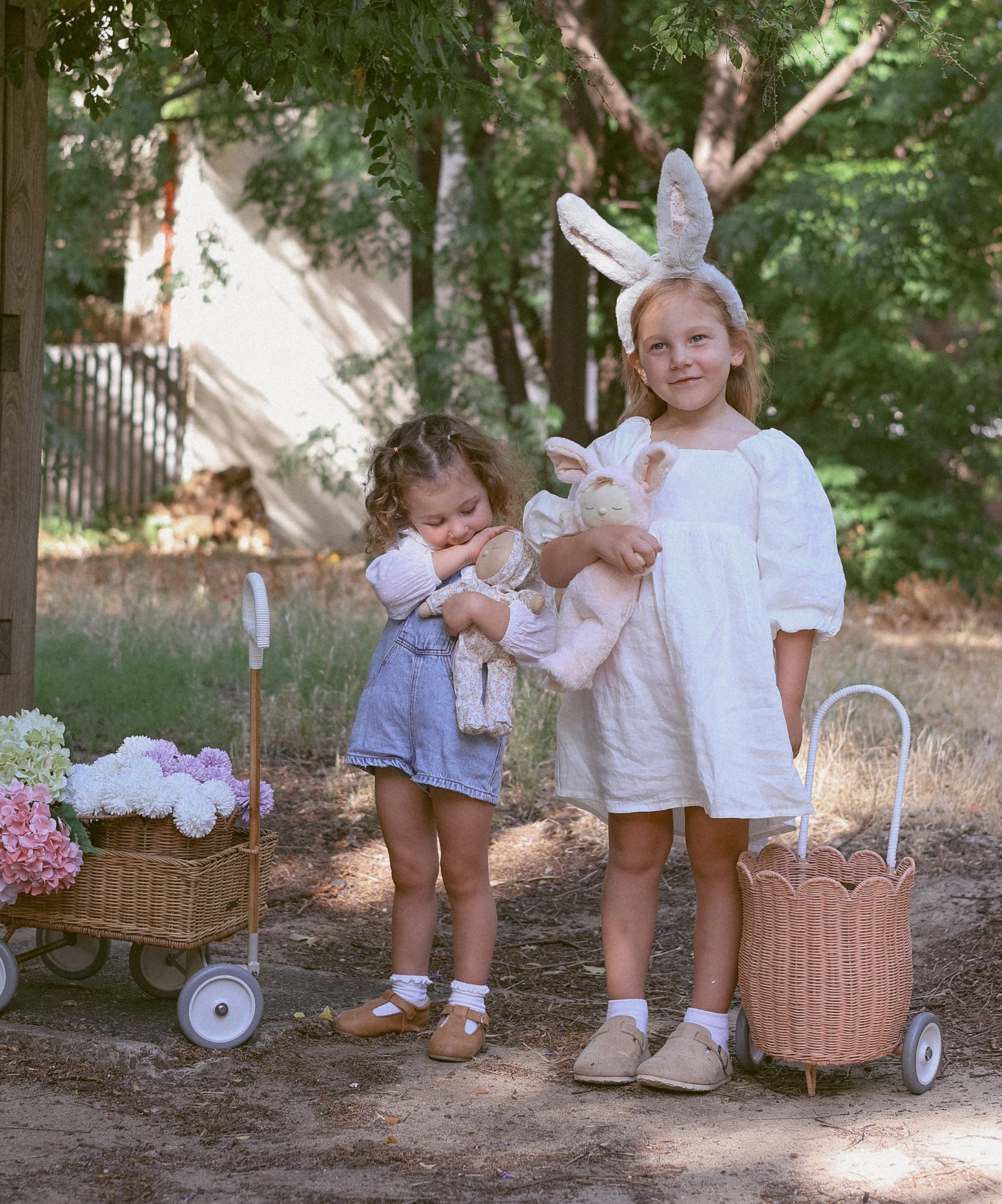 Two children standing outdoors. One is wearing the Olli Ella bunny ears headband and has a Seashell Pink rattan petal luggy by their side. Both children are holding Dozy Dinkum soft toys. There is also a rattan wagon off to the side.