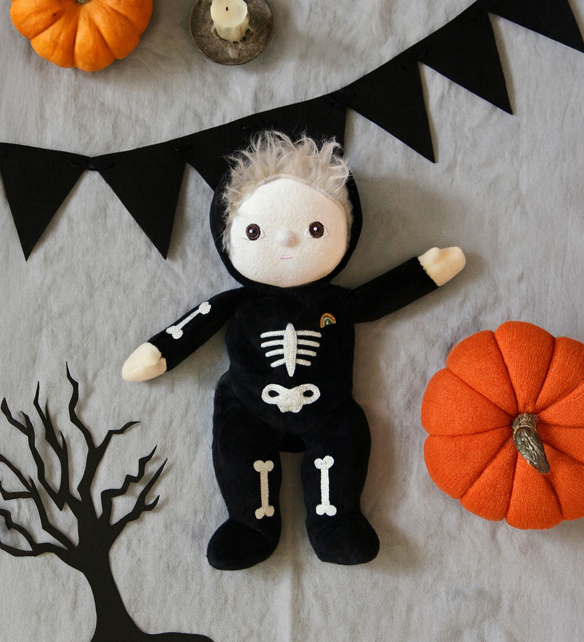 Olli Ella Scully Skeleton Dinky Dinkum Doll soft toy. The doll features glow in the dark bone details on the front along with a small embroidered rainbow on the chest. The toll has been placed in the middle of a Halloween themed flatlay. These soft toys are part of Olli Ella's Halloween collection available here at Babipur.