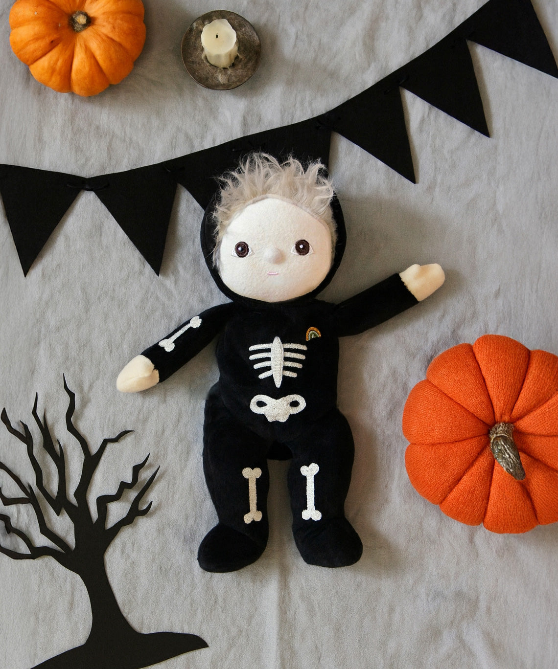 Olli Ella Scully Skeleton Dinky Dinkum Doll soft toy. The doll features glow in the dark bone details on the front along with a small embroidered rainbow on the chest. The toll has been placed in the middle of a Halloween themed flatlay. These soft toys are part of Olli Ella's Halloween collection available here at Babipur.