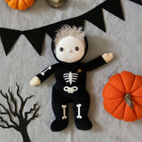 Olli Ella Scully Skeleton Dinky Dinkum Doll soft toy. The doll features glow in the dark bone details on the front along with a small embroidered rainbow on the chest. The toll has been placed in the middle of a Halloween themed flatlay. These soft toys are part of Olli Ella's Halloween collection available here at Babipur.