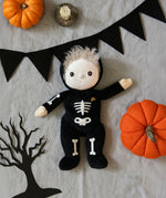 Olli Ella Scully Skeleton Dinky Dinkum Doll soft toy. The doll features glow in the dark bone details on the front along with a small embroidered rainbow on the chest. The toll has been placed in the middle of a Halloween themed flatlay. These soft toys are part of Olli Ella's Halloween collection available here at Babipur.